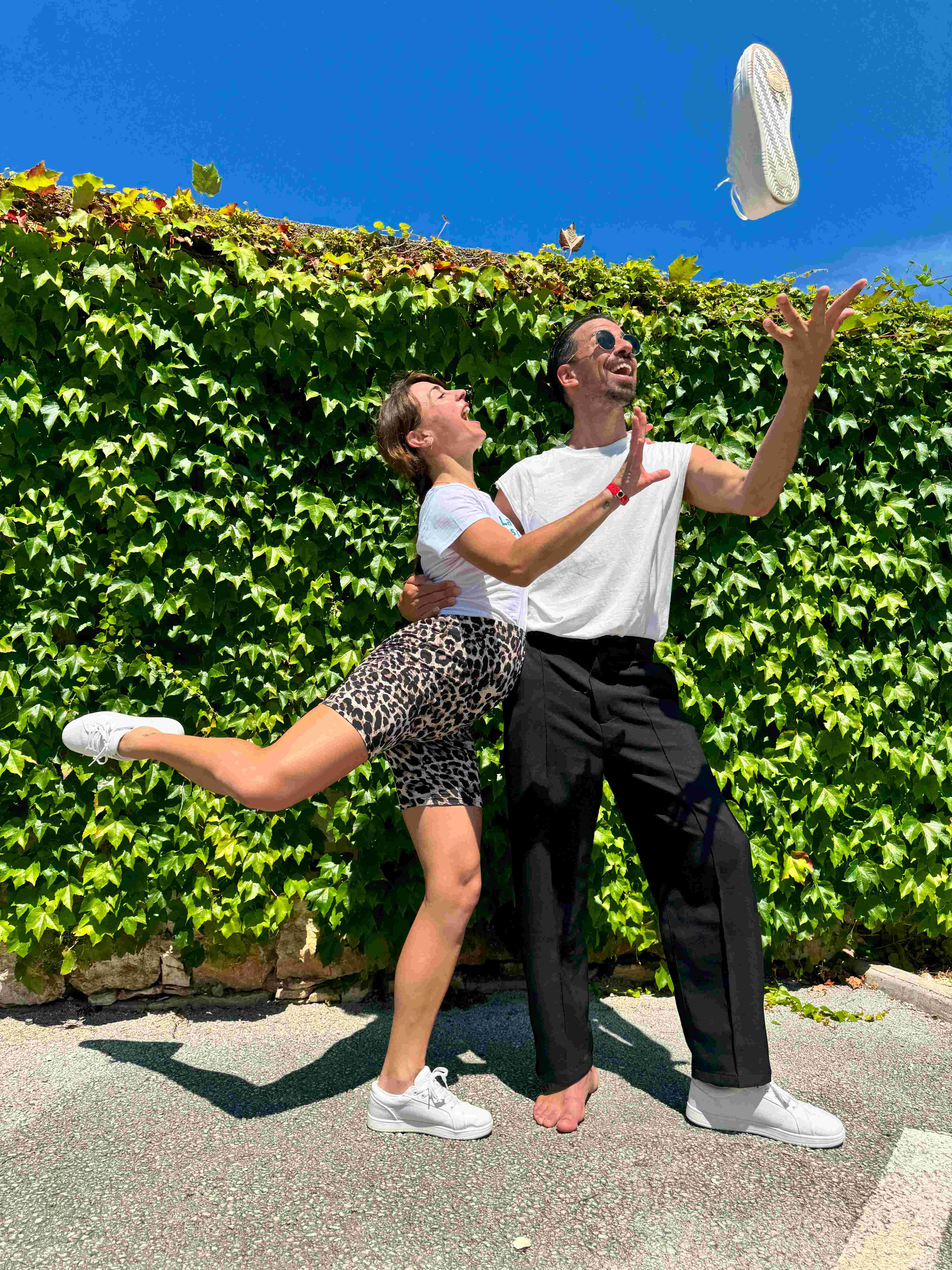 Ein Paar posiert freudig vor Efeu und wirft an einem sonnigen Tag spielerisch einen Schuh in die Luft.